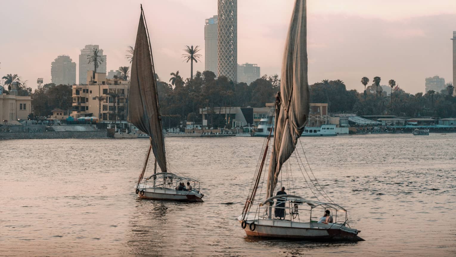 Two felucca boats drift on the water in the evening light, a busy shoreline and Cairo Tower in the distance. 
