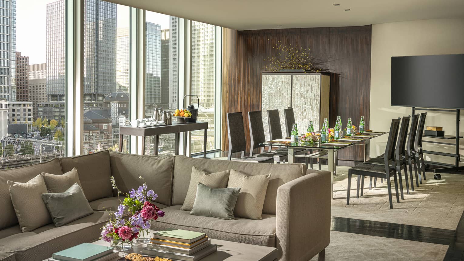 Living area with a large window, city skyline view, beige sectional sofa, table with refreshments, dining area with black chairs and home theatre