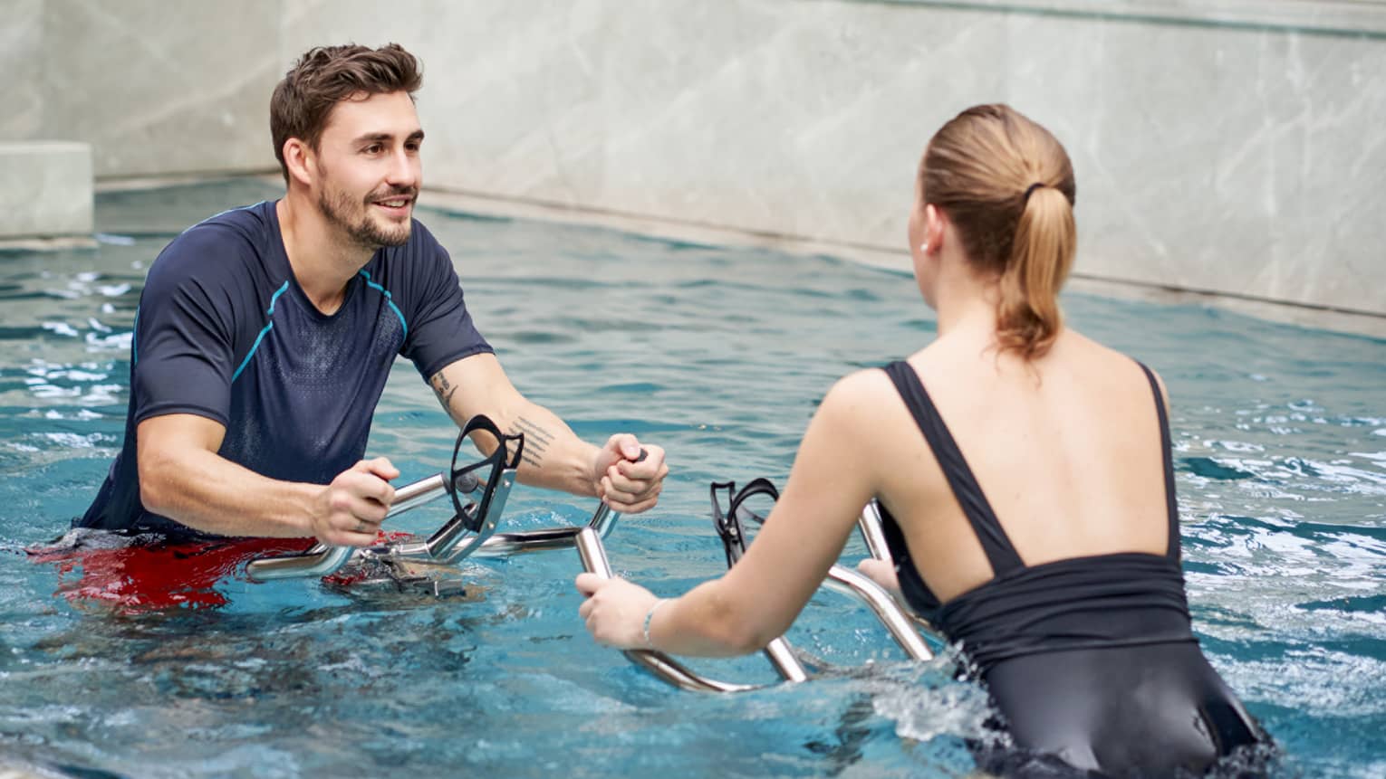 A trainer and a guest face each other in a marble-walled indoor pool as they exercise on underwater stationary bicycles.