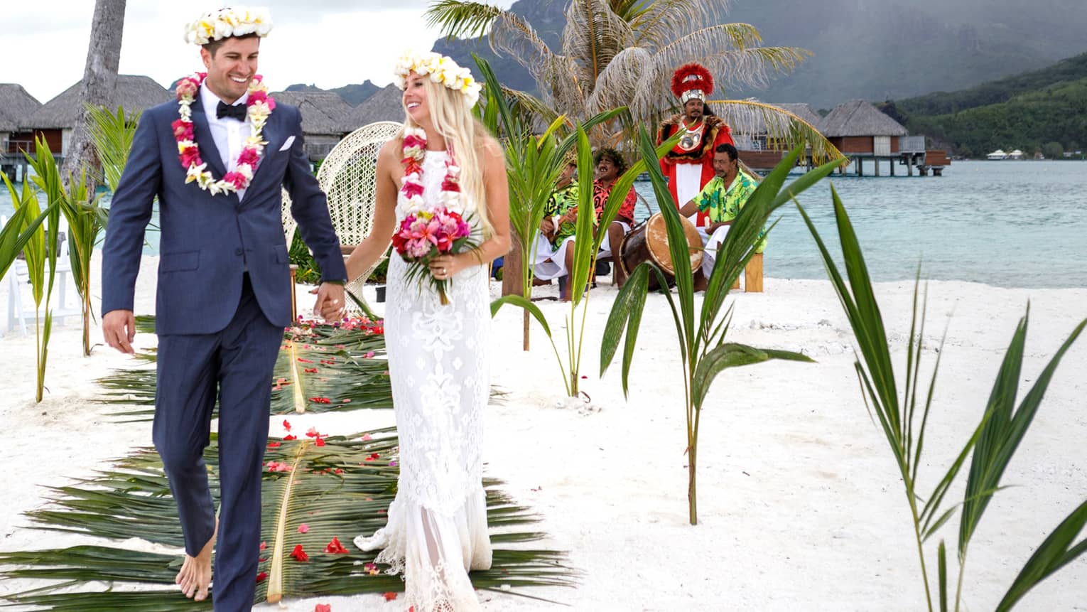 Bride and groom wearing floral crowns walk down aisle on white sand beach with palms