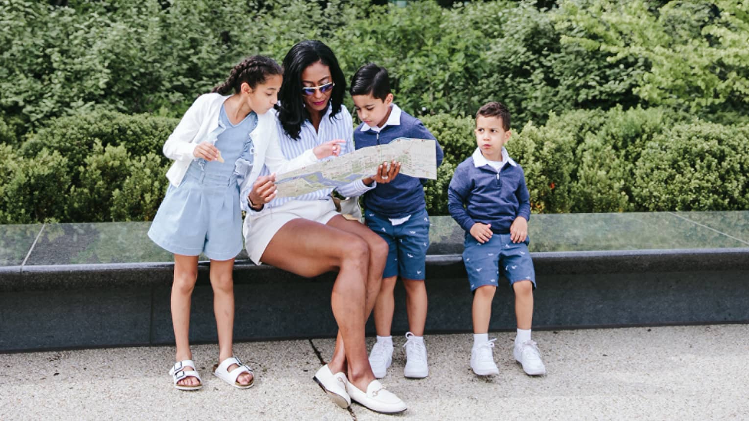 Woman and three young children sit on bench in city with landscaping behind them
