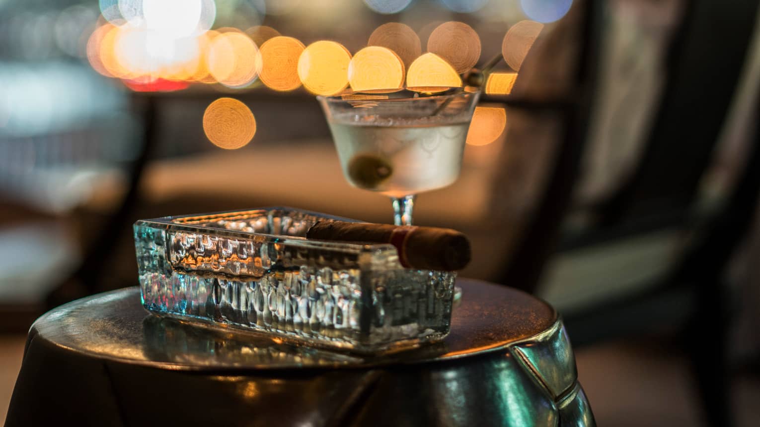 Cigar in heavy rectangular glass ashtray beside martini glass with olive 