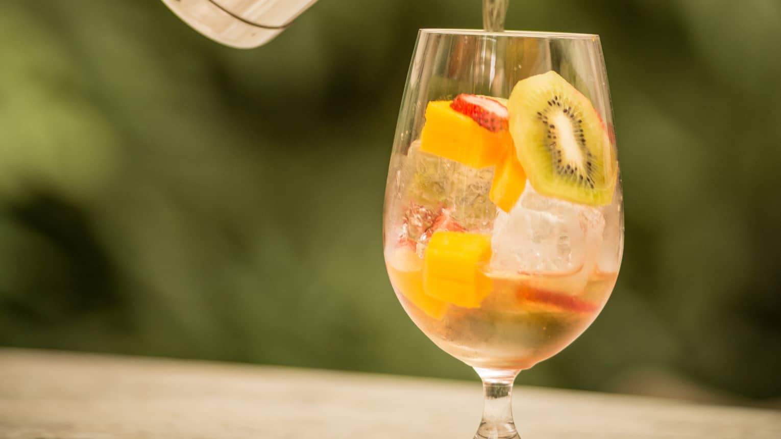 A person mixes a drink in a large water glass containing chopped kiwi, strawberry and mango.