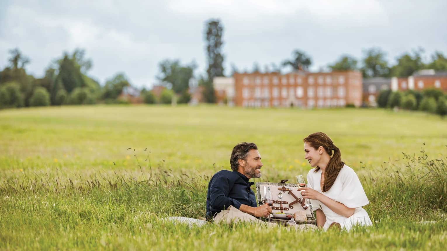 Couple with wine glasses lies in tall green grass with open picnic basket, hotel manor in background