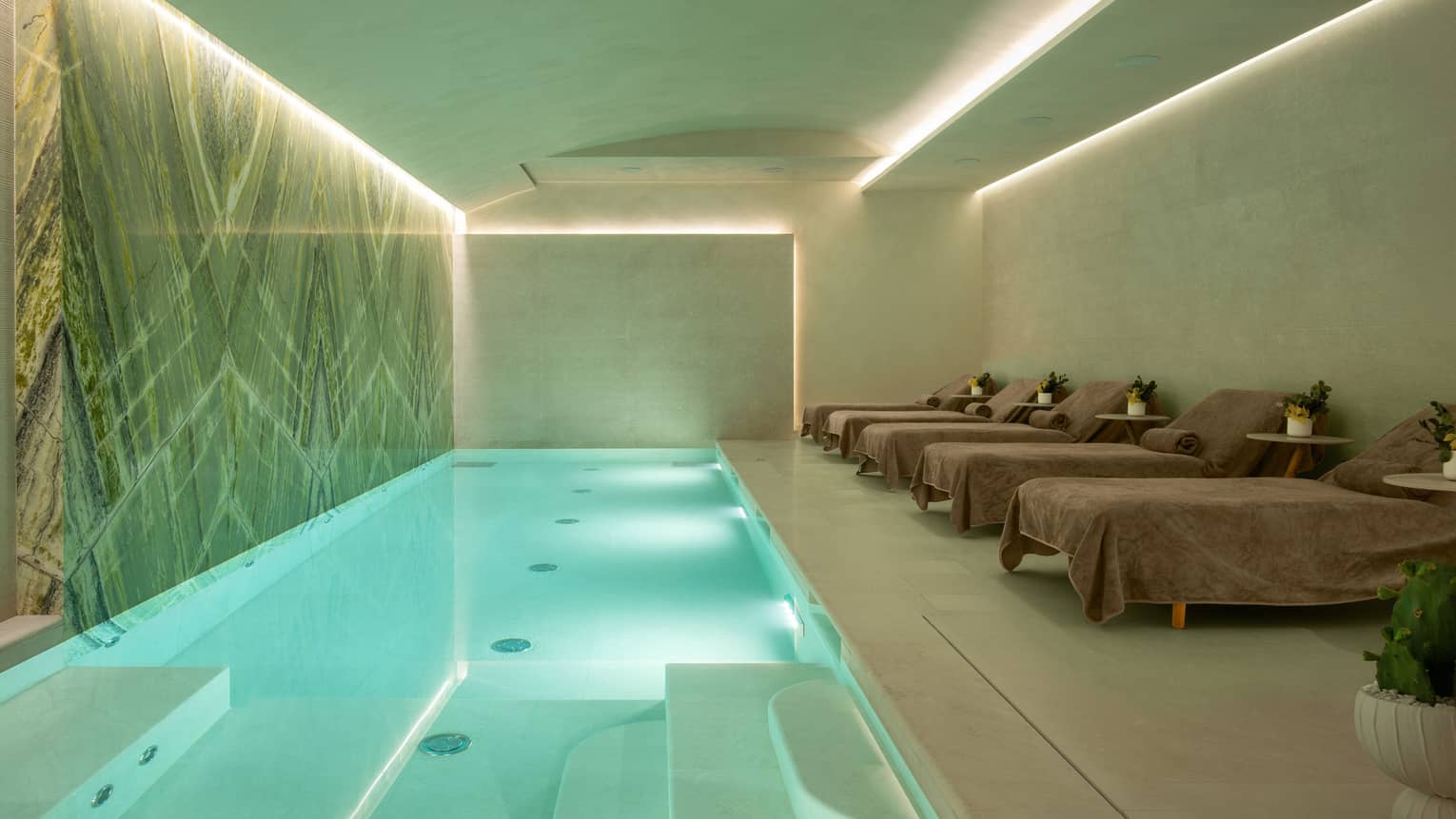 Uplit rectangular indoor pool flanked by marble wall and row of lounge chairs