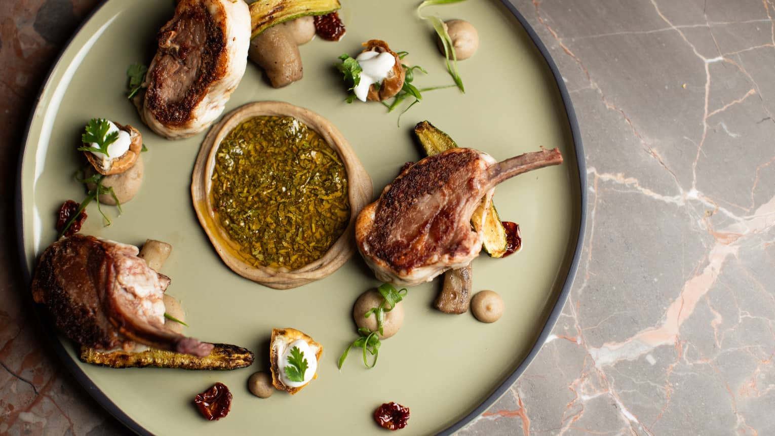 Three grilled lamb chops plated with eggplant and an assorment of garnishes on a large, round, light-green plate