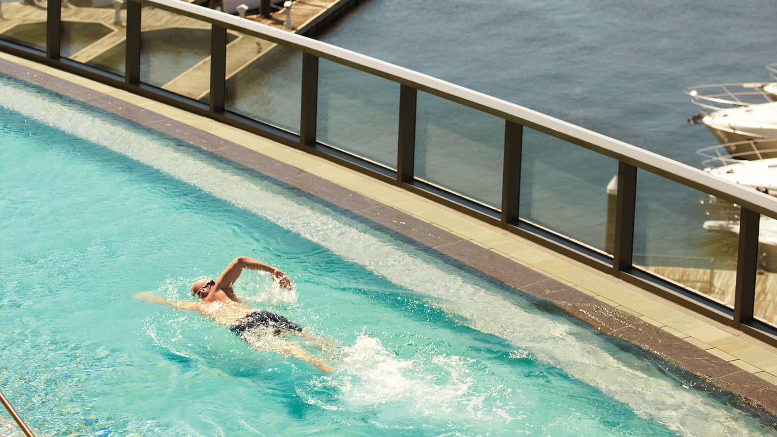 Man in swimsuit, goggle swims laps in outdoor pool with glass panels overlooking harbour