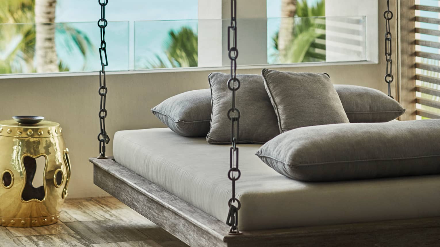 Hanging daybed with gray cushions on a resort balcony, accompanied by a gold decorative side table, overlooking tropical palm trees and the sea