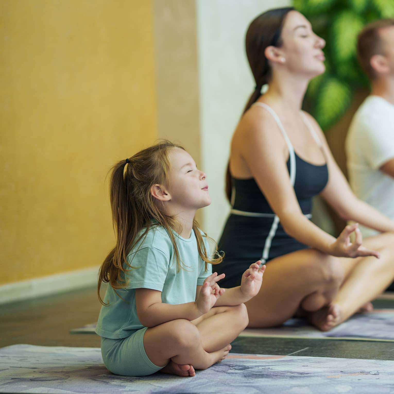 Two adults and one child each sit on a yoga mat with their legs crossed, eyes closed and hands held on their knees