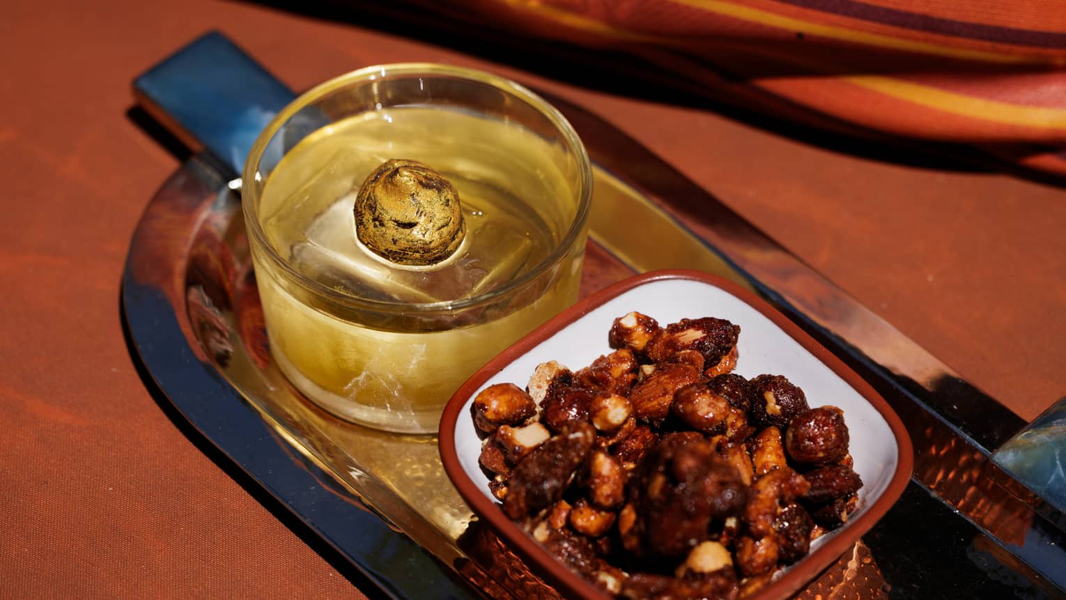 Whiskey-based cocktail in a rocks glass paired with a bowl of nuts on a curved rectangular golden serving tray