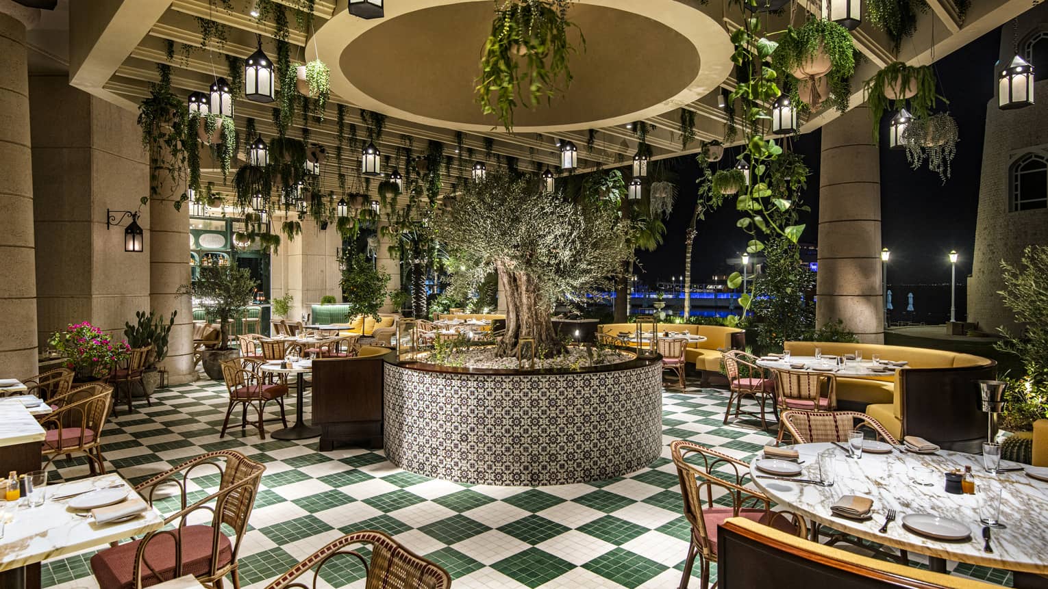 Curiosa restaurant interior featuring green-and-white checkerboard tile floors, rattan chairs, yellow banquets and plants and lanterns hanging from the ceiling
