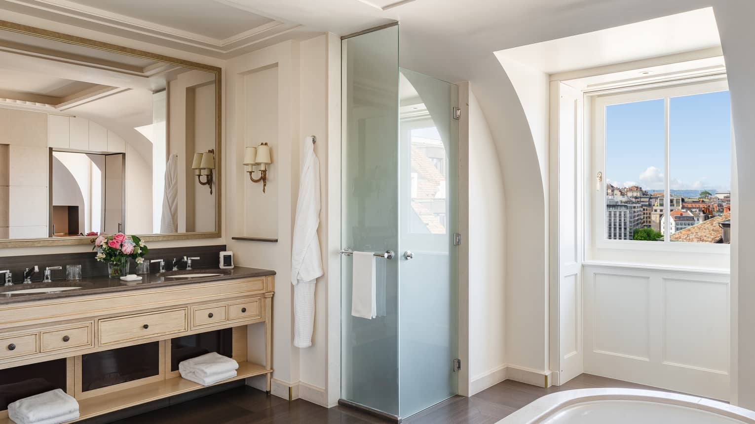 Luxury hotel bathroom with a large mirror, double sink vanity, glass shower door and a window offering city view