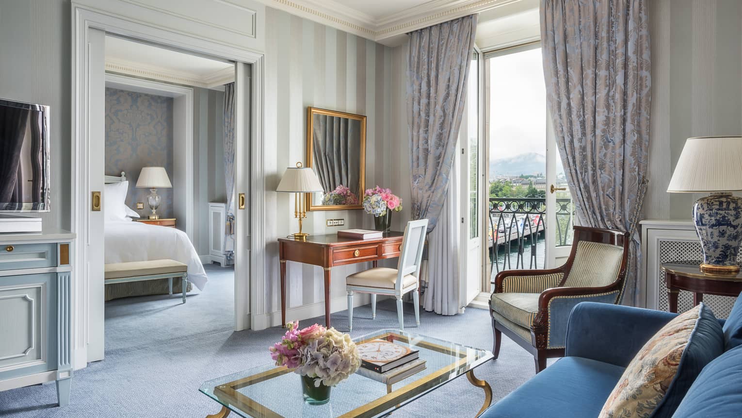 Luxury hotel suite living area with a blue sofa, glass coffee table, desk and armchair by a window with a city view and a separate bedroom