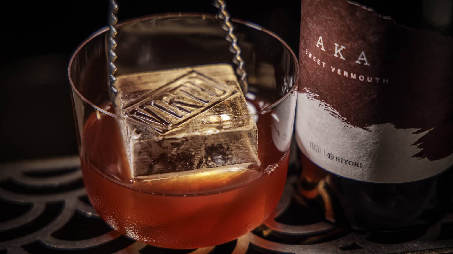 A pair of metal tongs lowers a large square ice cube into a chilled cocktail next to a bottle of Vermouth
