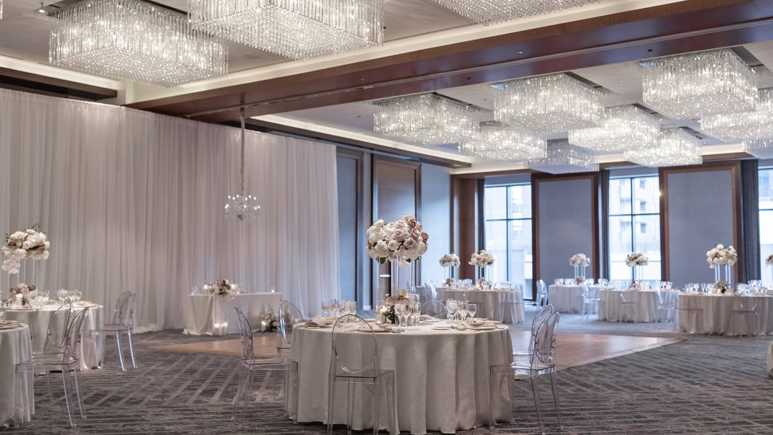 A ballroom with round tables with flower centerpieces surrounding a section of hardwood floor, many lights hang from the ceiling.