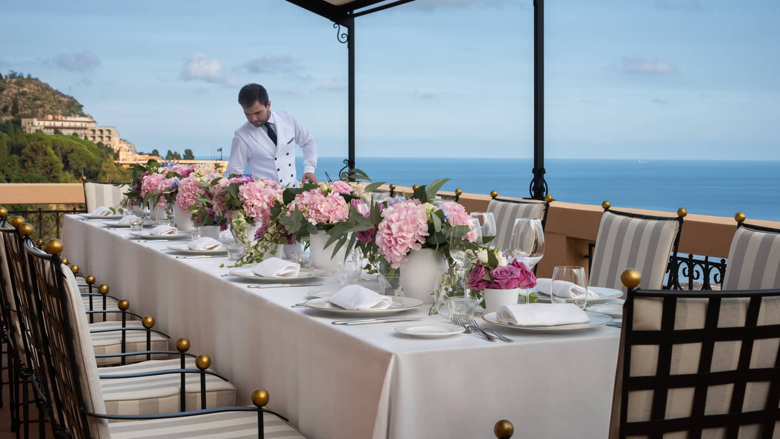Male server sets long, private table on outdoor terrace overlooking sea