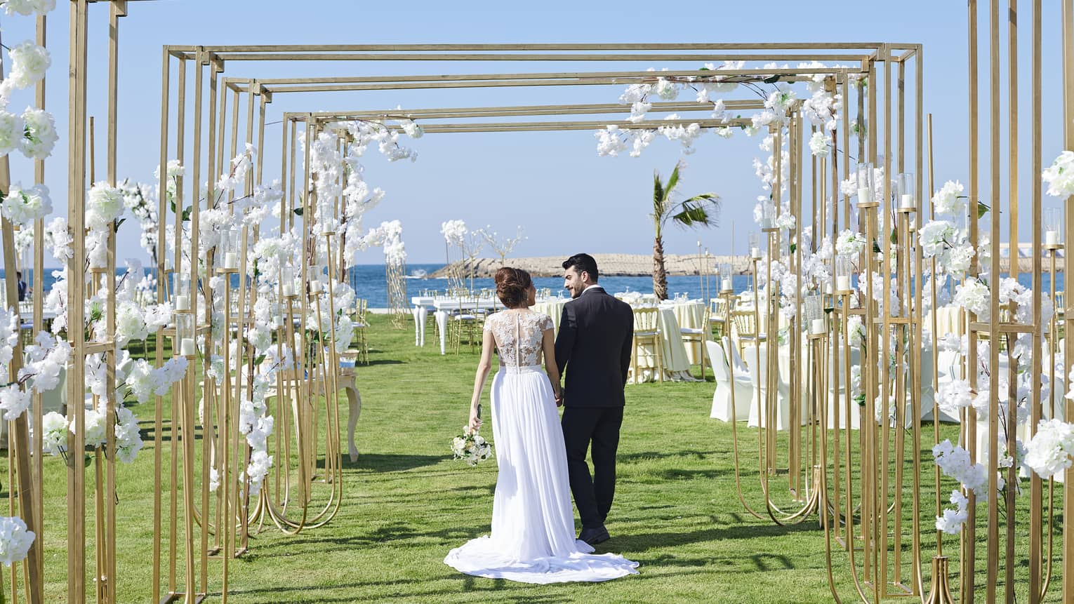 Rear view of wedding couple holding hands and smiling under a floral and gold archway path on lawn