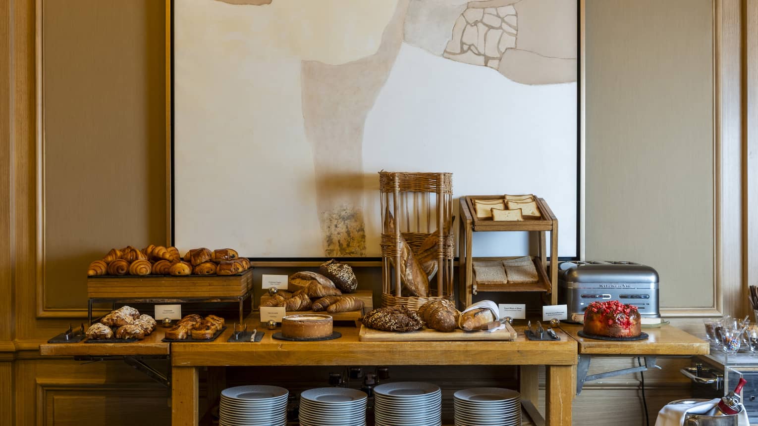 A wood sideboard display of breakfast food items in front of a beige wall with a piece of abstract art on it