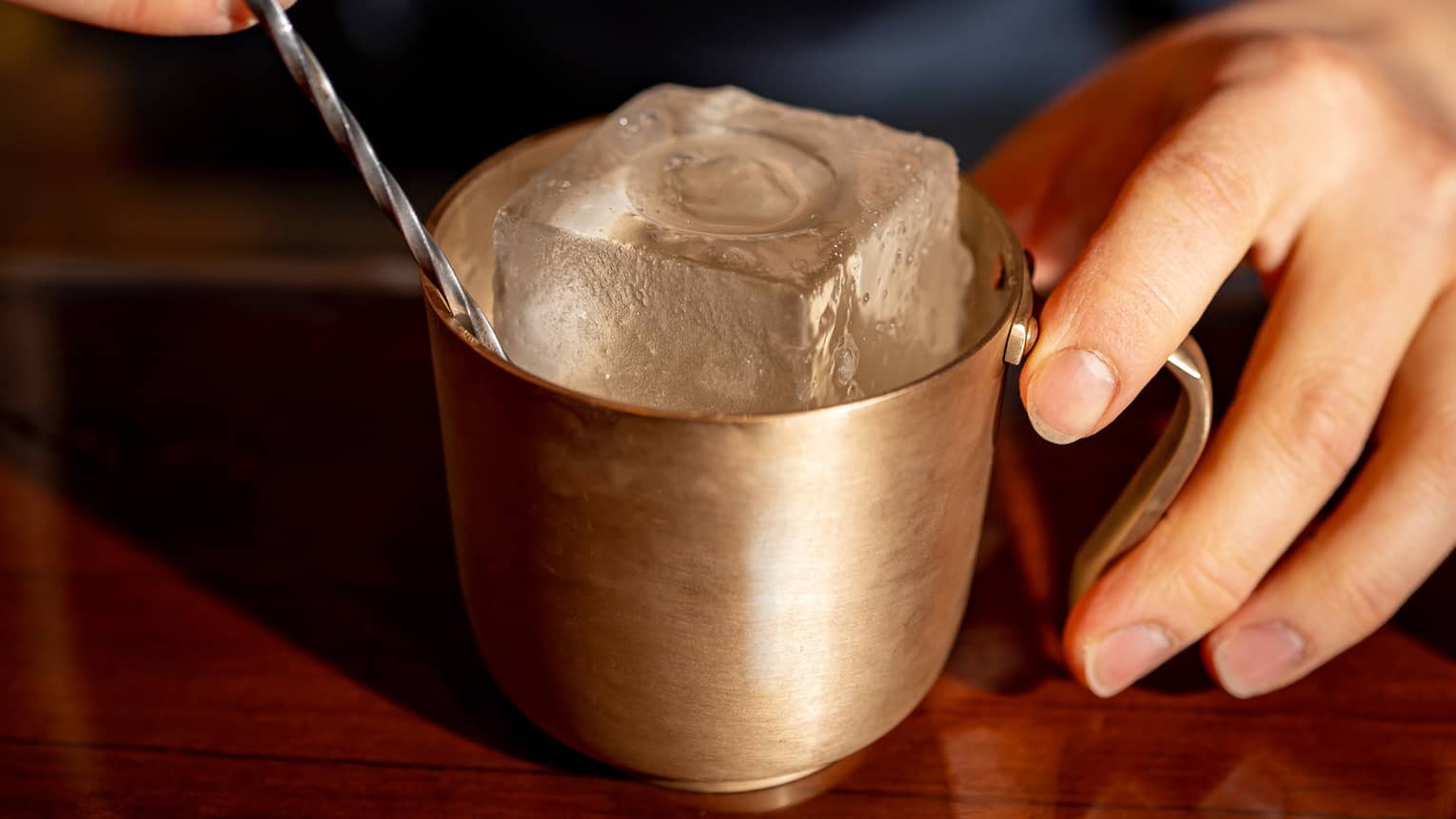 Close-up of a hand on a gold metallic mug containing a giant embossed ice cube and a twisting metal stir stick.