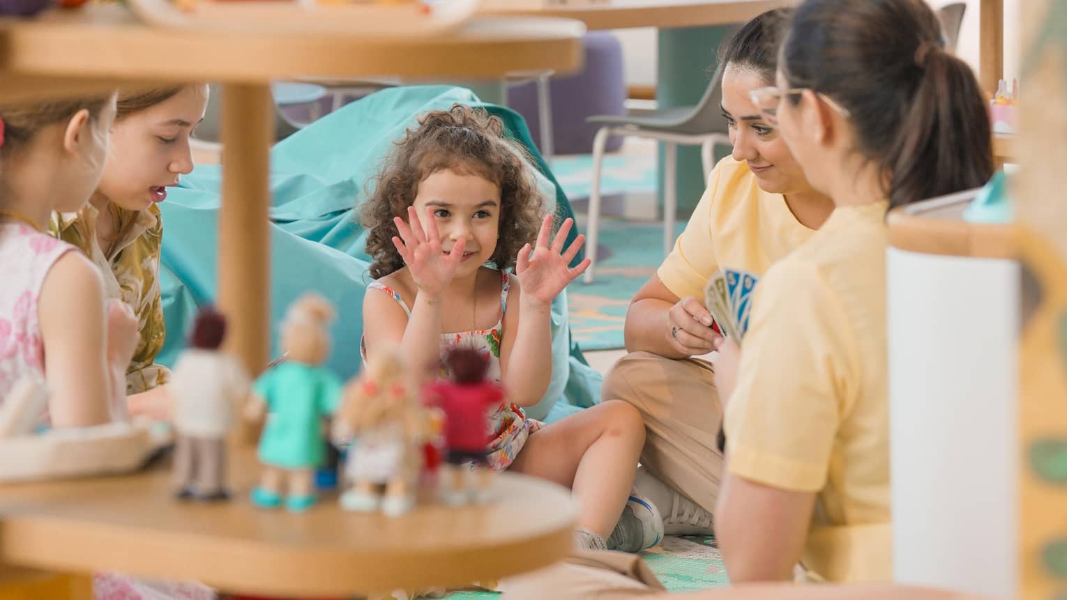 Three young children sit on the floor in a playroom playing cards with two adults
