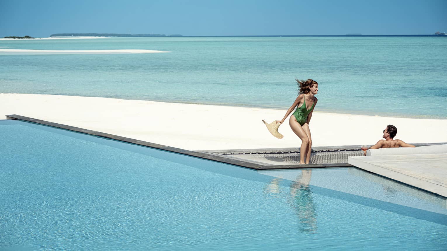 Woman and green one-piece swimsuit relaxes poolside with her male partner in the infinity pool, the vast blue waters of the lagoon behind them