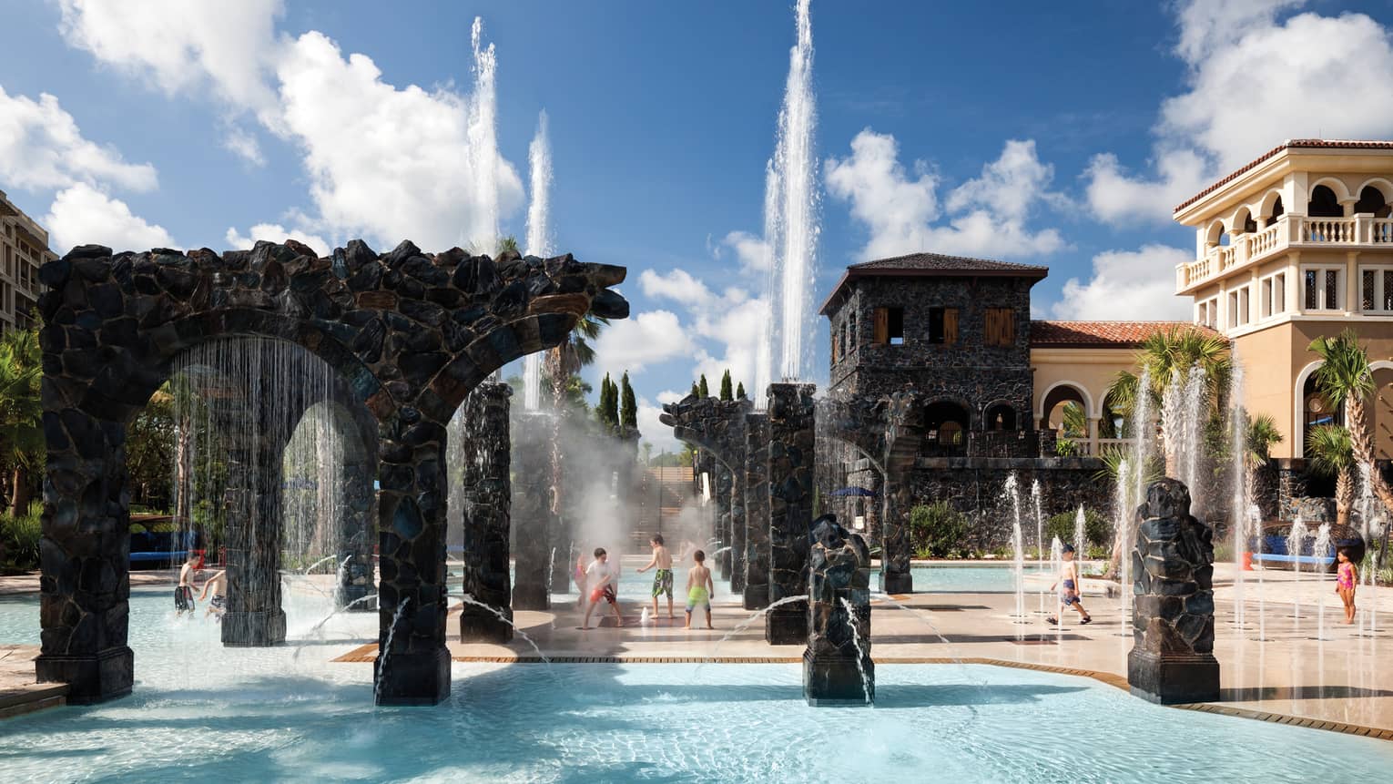 Kids play in water, under sprinklers and stone pillars in the Splash Zone water park