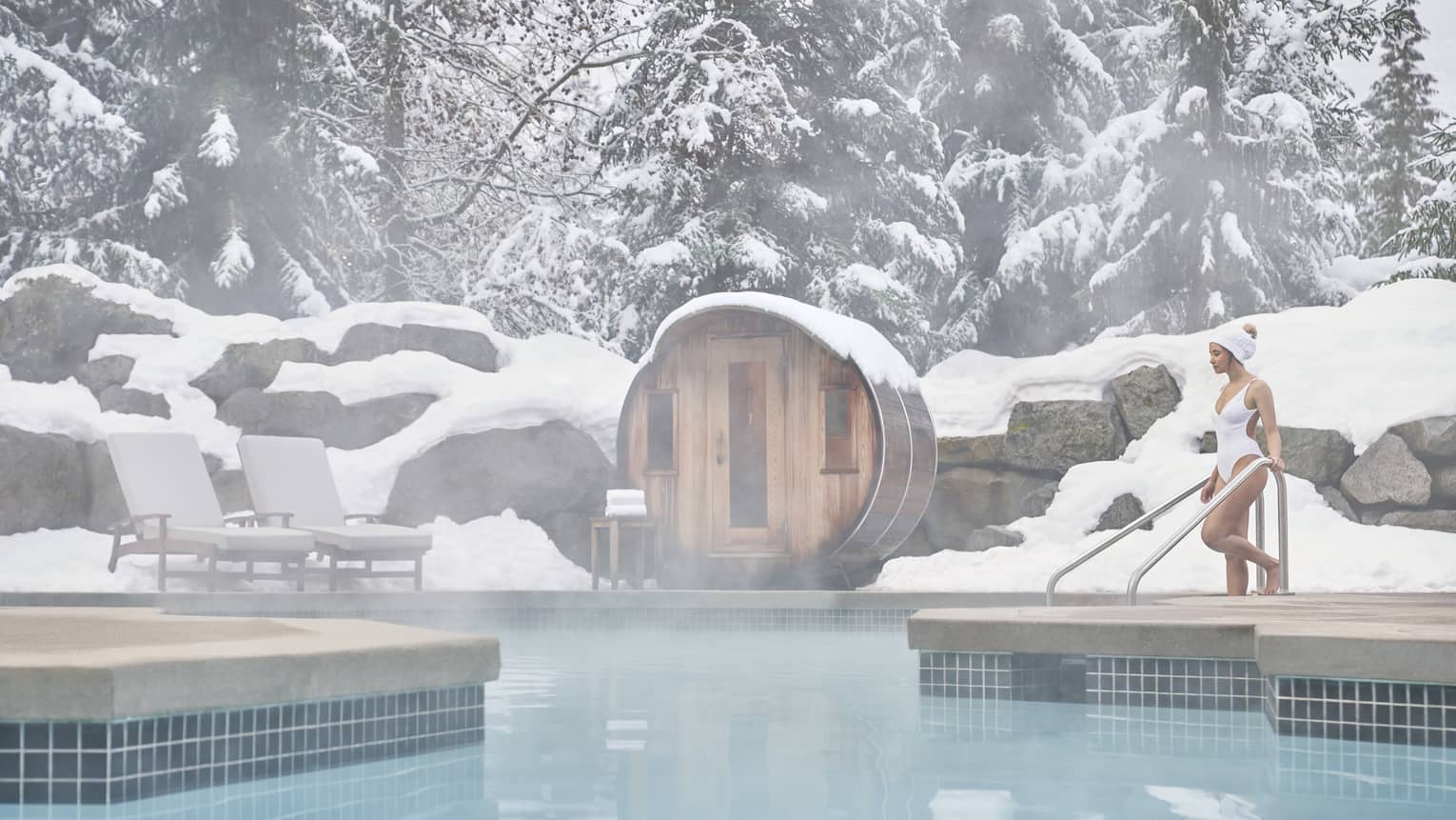 A woman in a white swimsuit enters the outdoor heated pool as steam rises around her. She is surrounded by snow-capped trees.