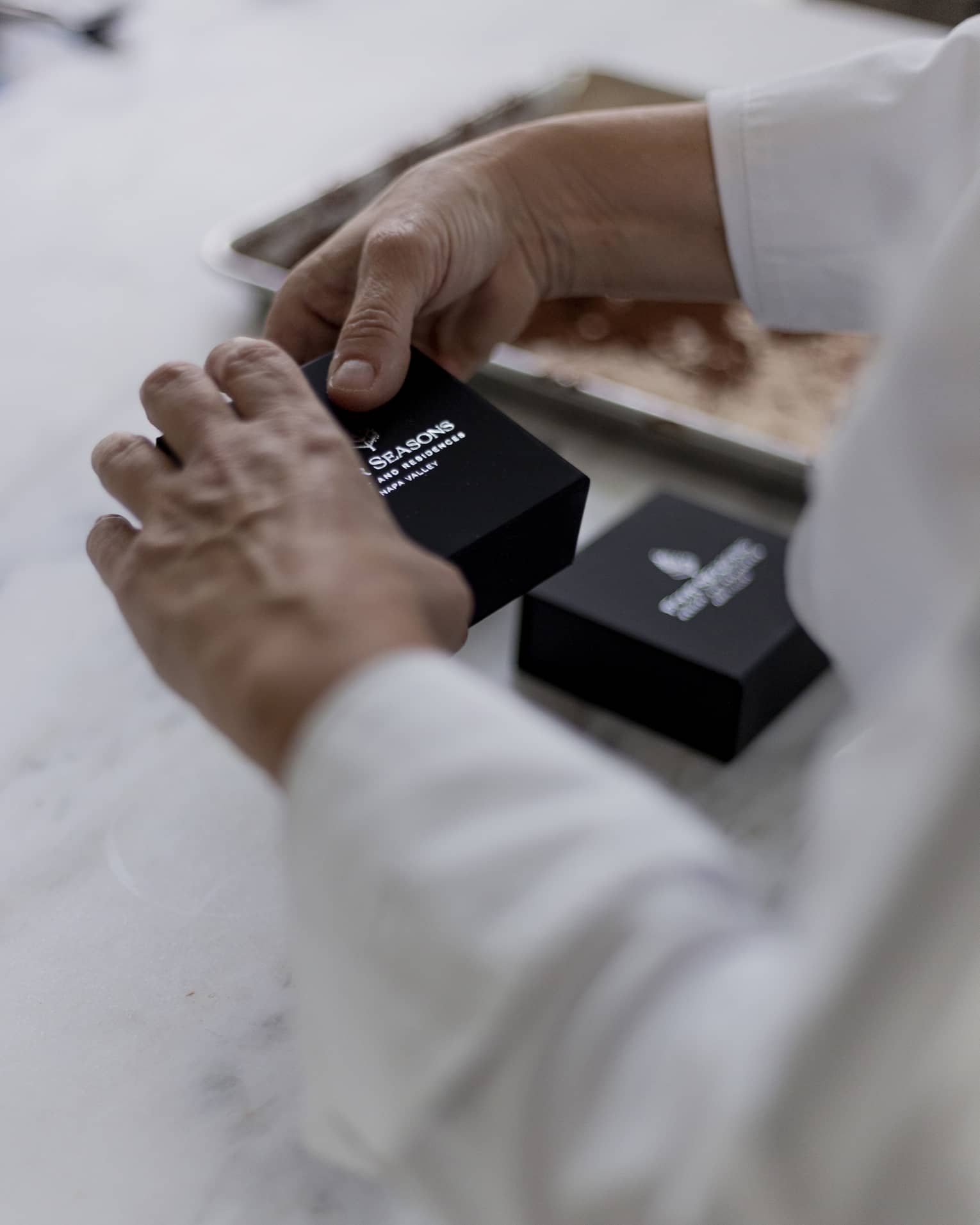 Hands holding a small, black box with the Four Seasons name and logo in silver; a blurred cookie sheet in the background.