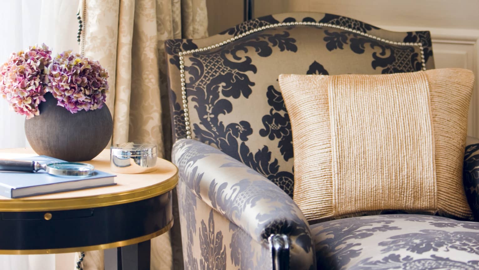 Close-up of armchair with black-and-cream Damask print, gold accent pillow, table with fresh purple flowers in black vase