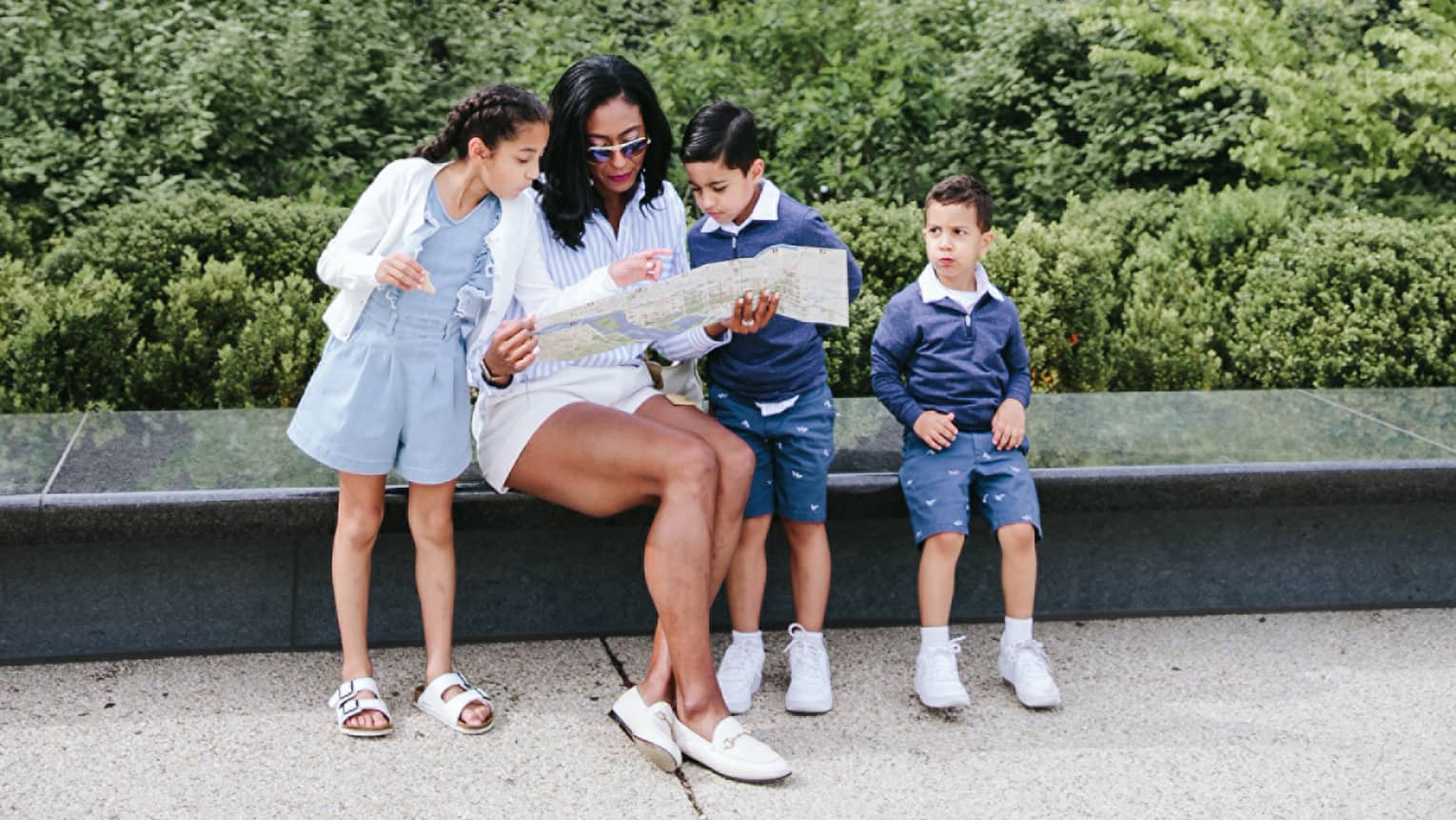 Woman and three young children sit on bench in city with landscaping behind them