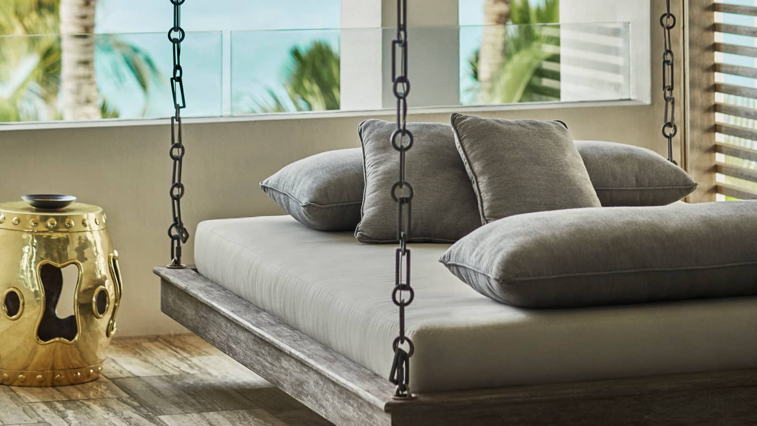 Hanging daybed with gray cushions on a resort balcony, accompanied by a gold decorative side table, overlooking tropical palm trees and the sea