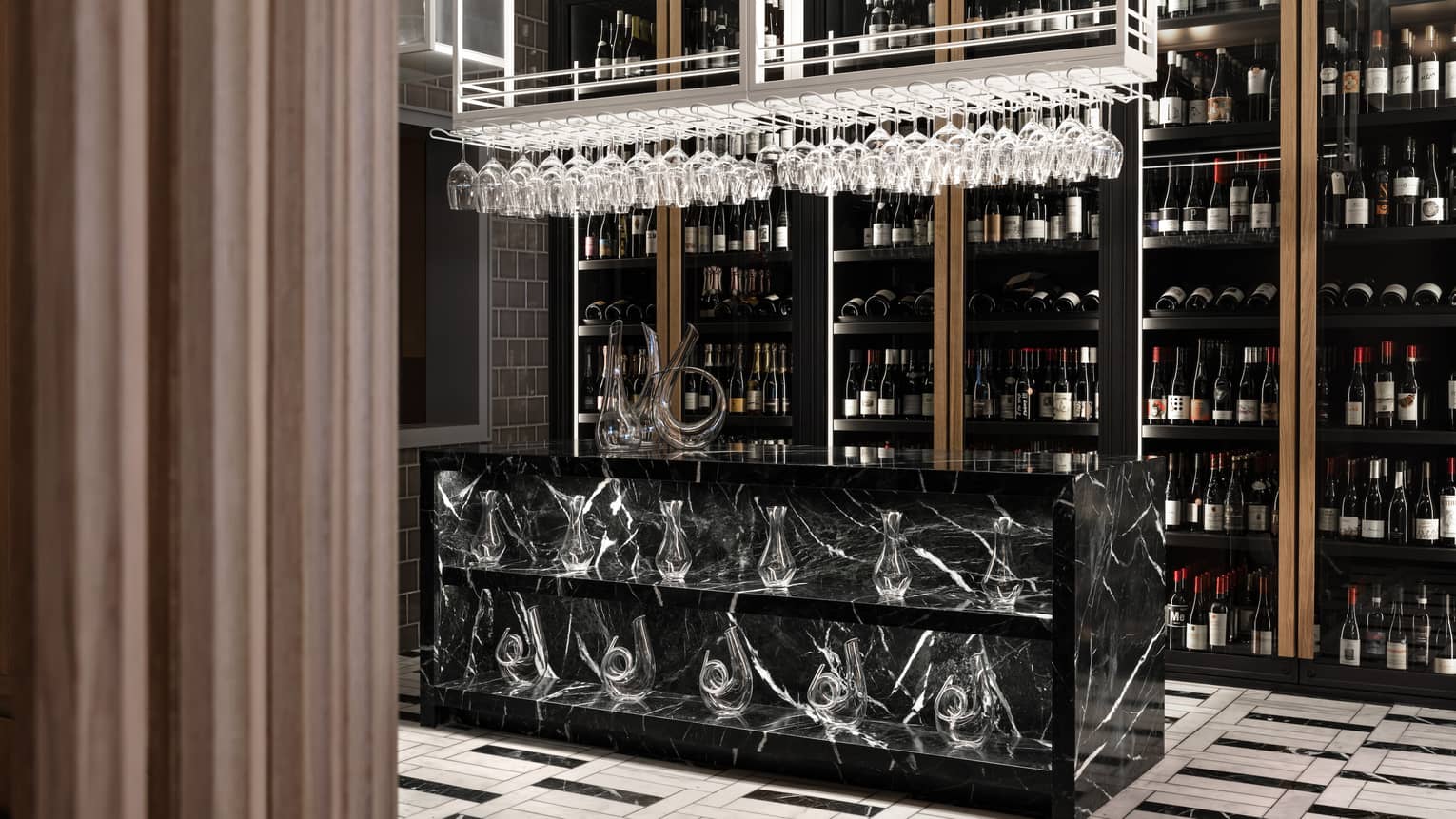 A wall lined from floor-to-ceiling with wine bottles sits behind a black marble counter that holds two shelves of glass decanters with hanging stemmed wine glasses hanging overhead