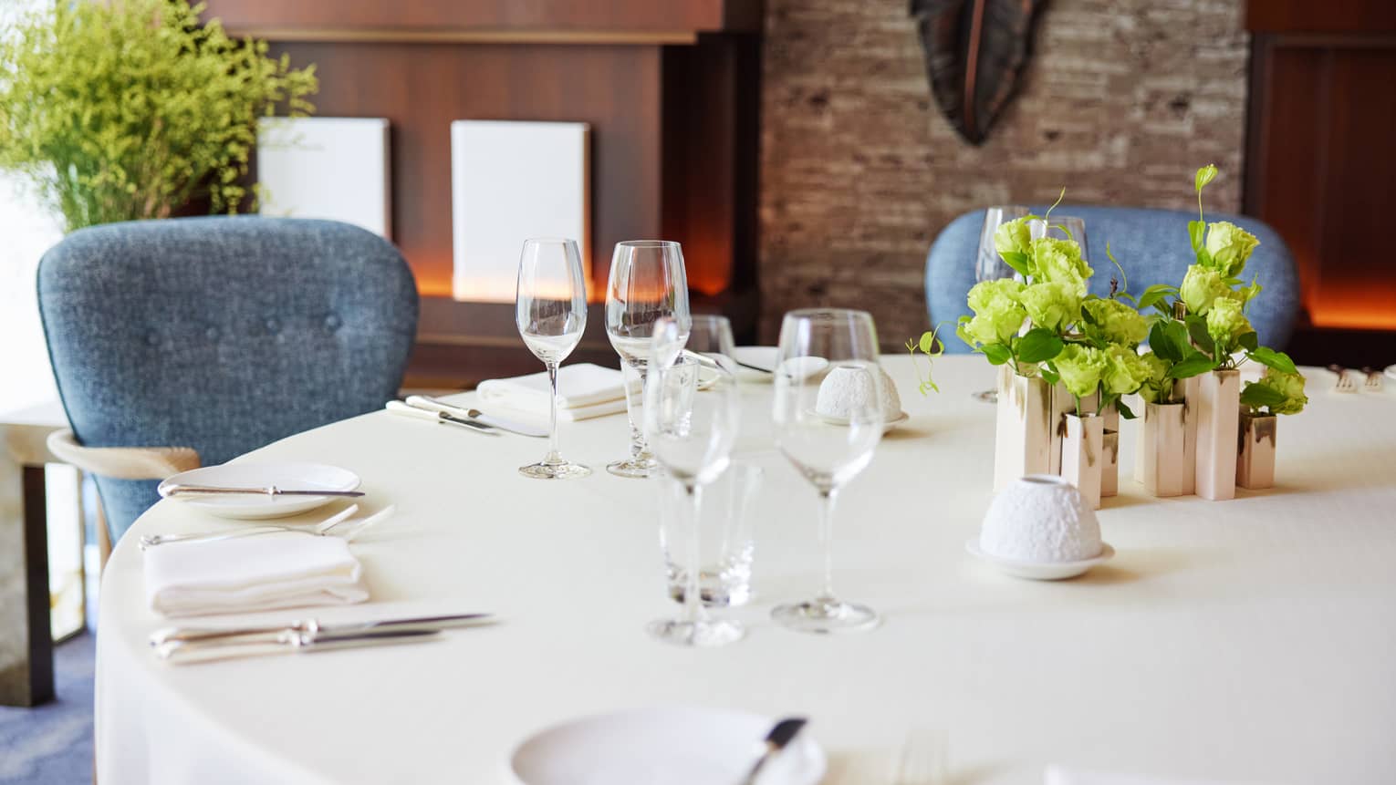Against a warm backdrop, round table set with white tablecloth, sparkling silver and wine glasses, fresh flowers at centre.