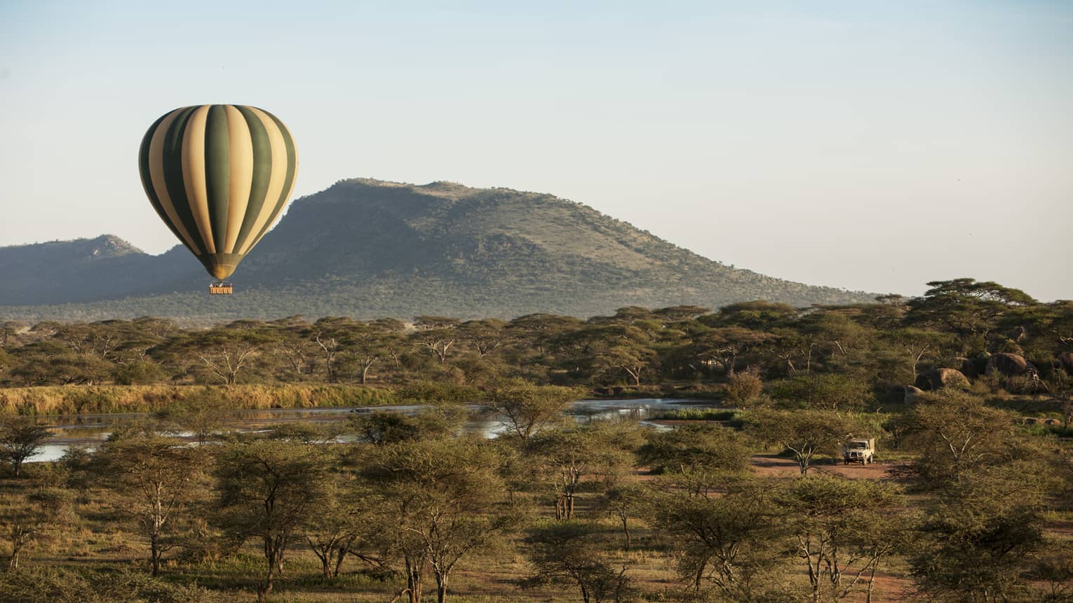 Stripped hot air balloon rising above Serengeti, trees and watering hole, mountain in background 