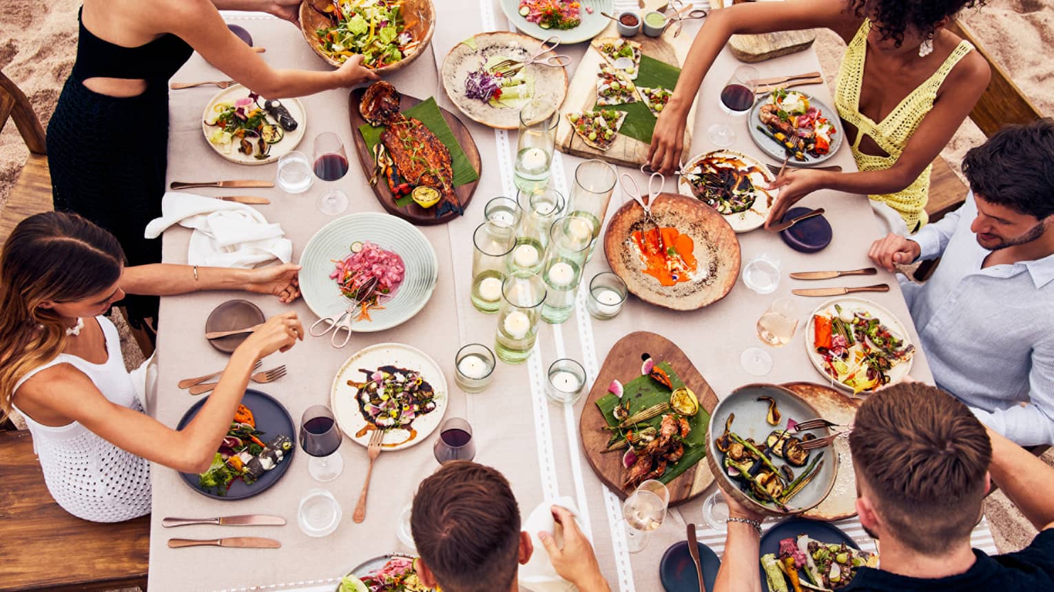 Beachside dining table with an assortment of vibrant dishes, candlelit glass holders and guests enjoying fresh seafood and grilled vegetables