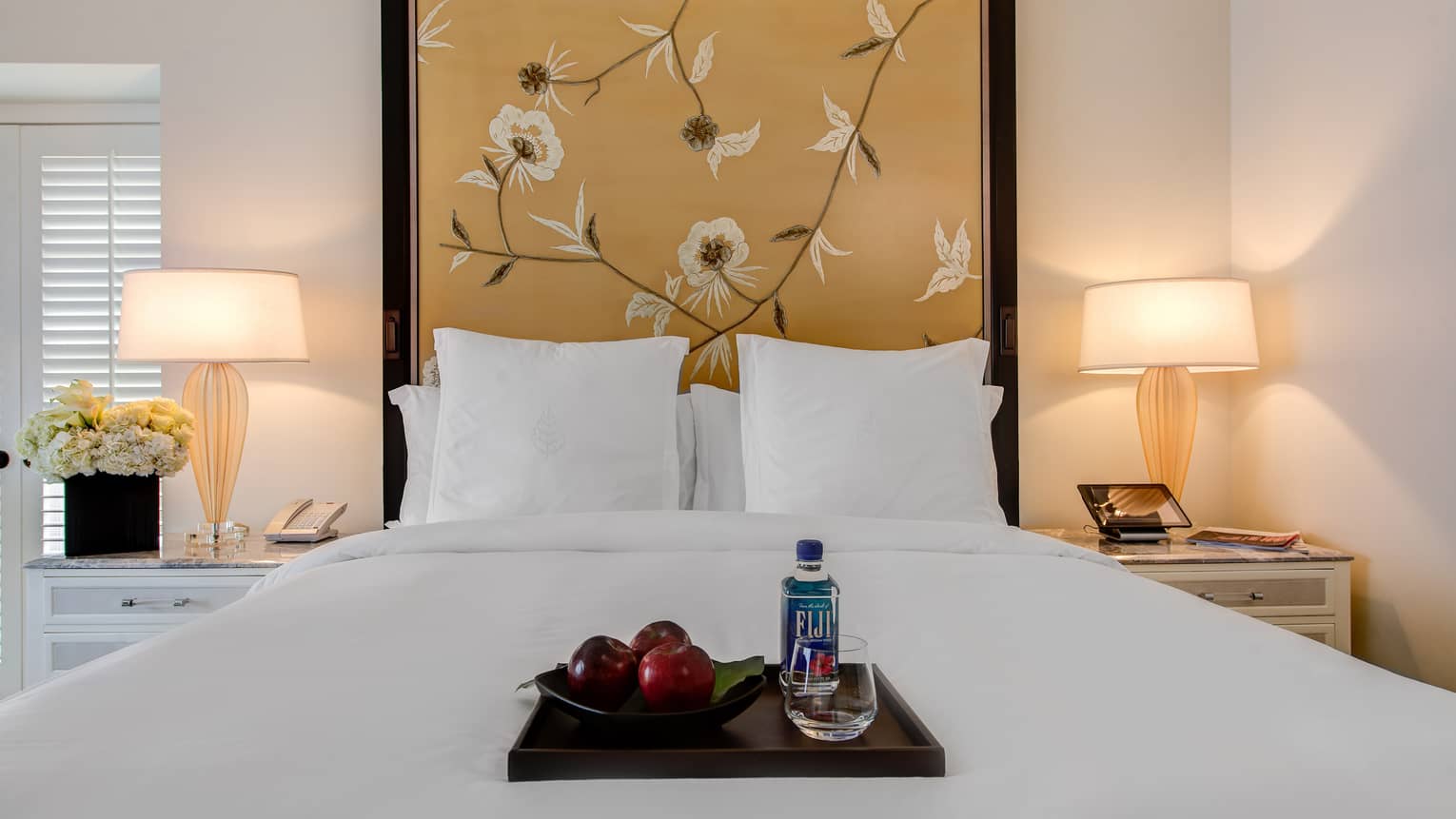 Deluxe Room hotel bed with tall floral headboard, wood tray with red apples, Fiji water, glass on bed