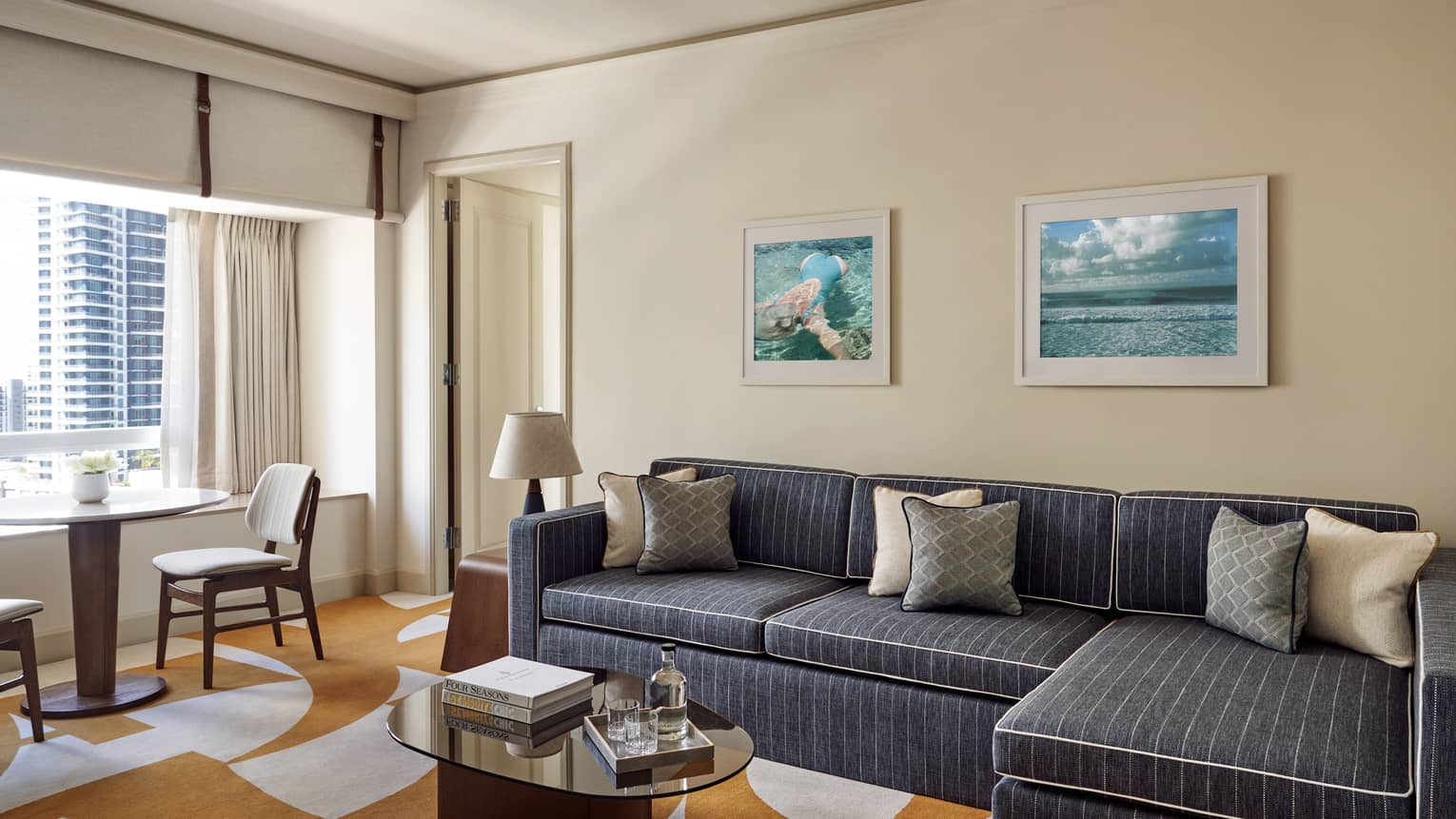 Hotel suite living room with dark grey sofa, amber and white abstract carpet, table and two chairs, window with city view