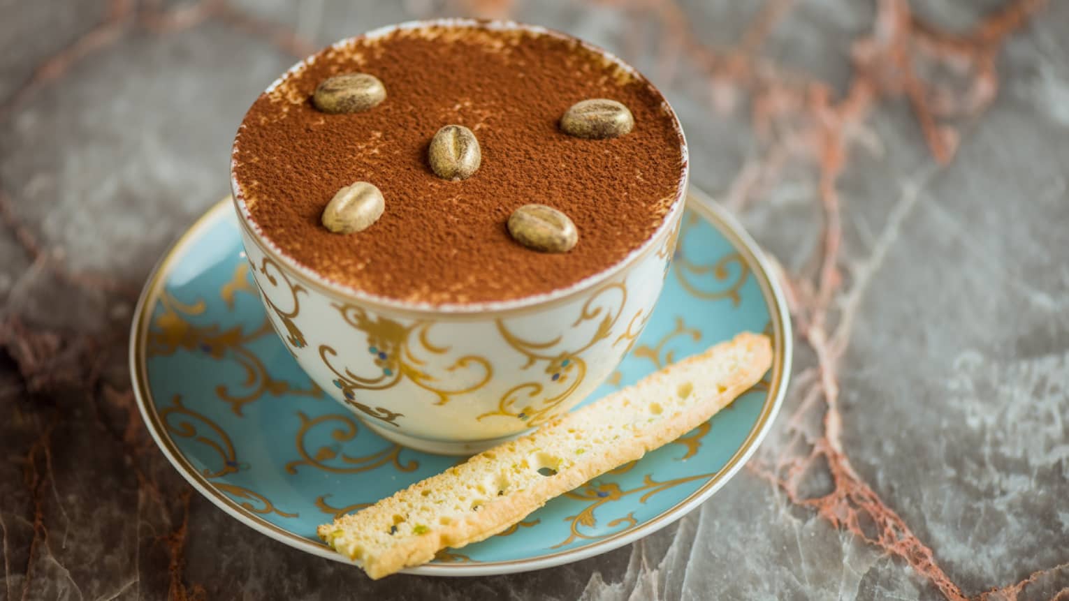 A cup of tiramisu topped with biscotti, served in a blue and gold teacup.