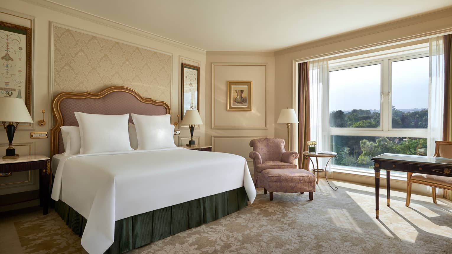 Deluxe Room bed with upholstered pink headboard, gold pattern wallpaper, pink silk armchair by sunny window