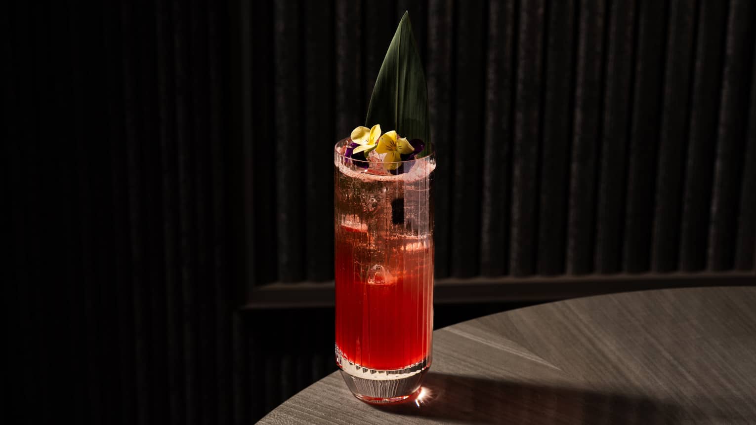 Red-hued cocktail garnished with yellow flowers served in a textured highball glass