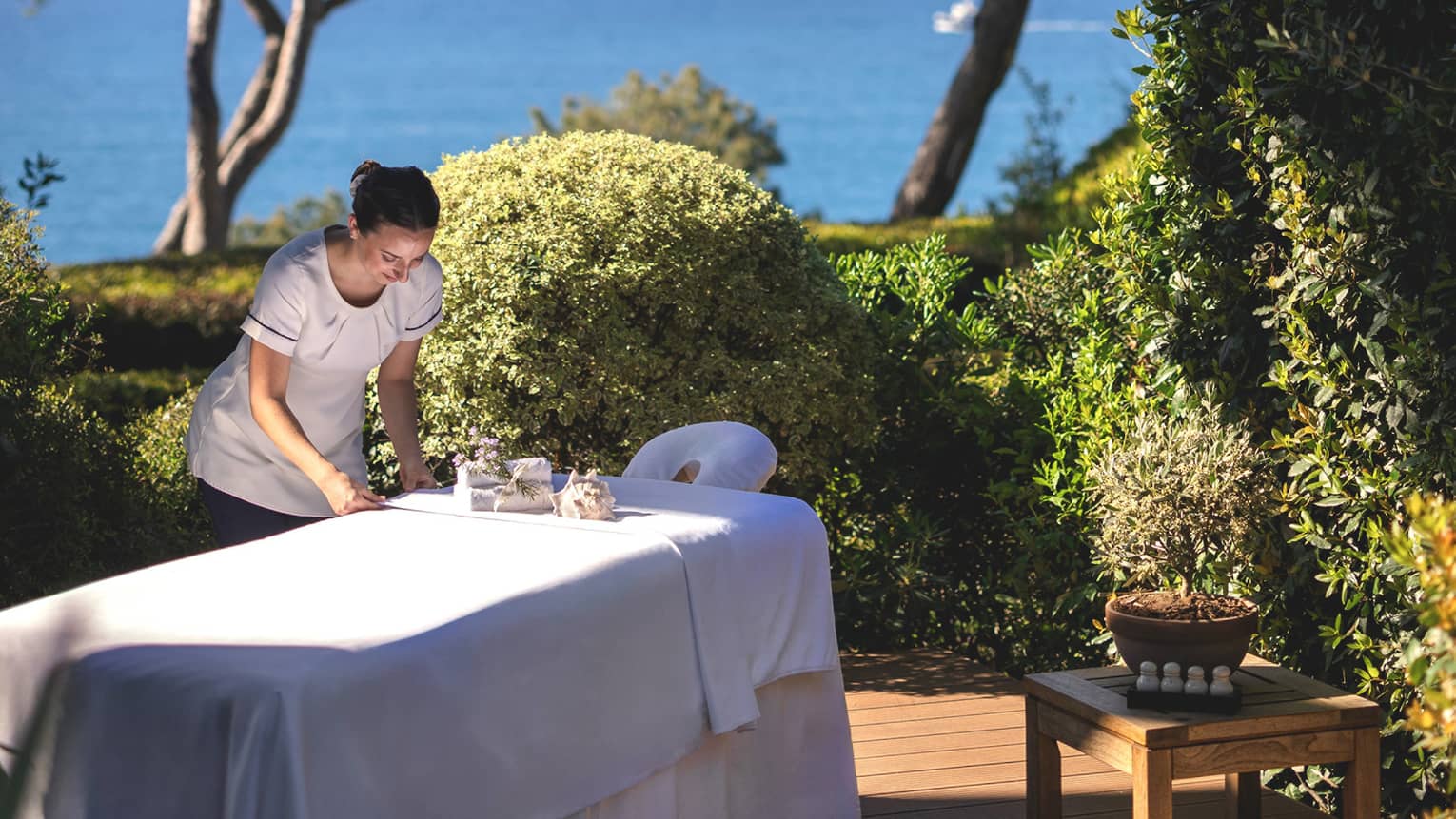 Spa therapist setting up spa table on outdoor platform surrounded by bushes and trees and overlooking the sea