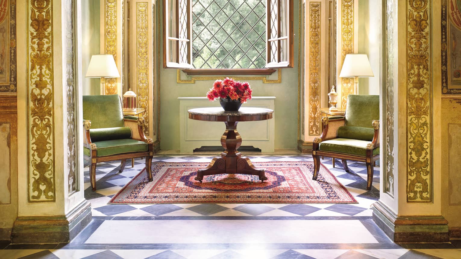 Green velvet armchairs around wood table with flowers, open window, gold ornate pillars