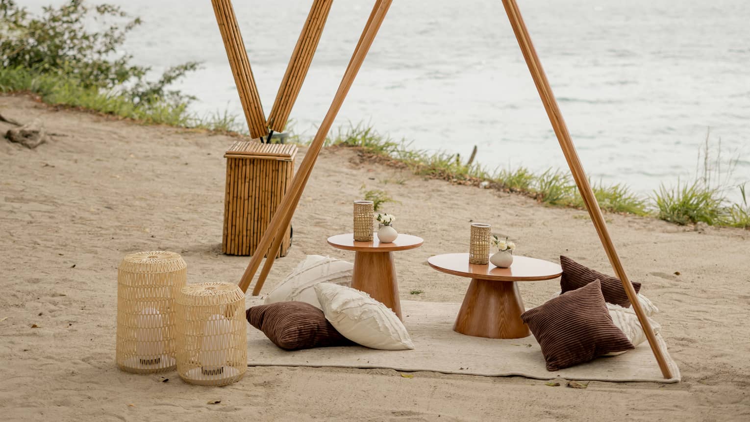 Rustic pergola with a blanket, pillows and a small table underneath set next to tiki torches and lanterns on a private beach