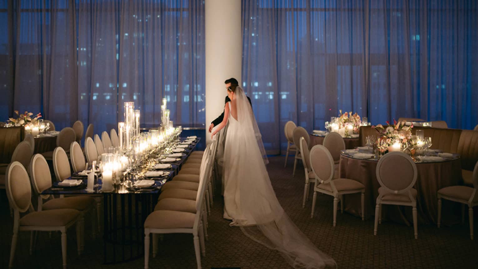 Wedding couple stands at classroom-style table in ballroom with floor-to-ceiling windows with sheer curtains