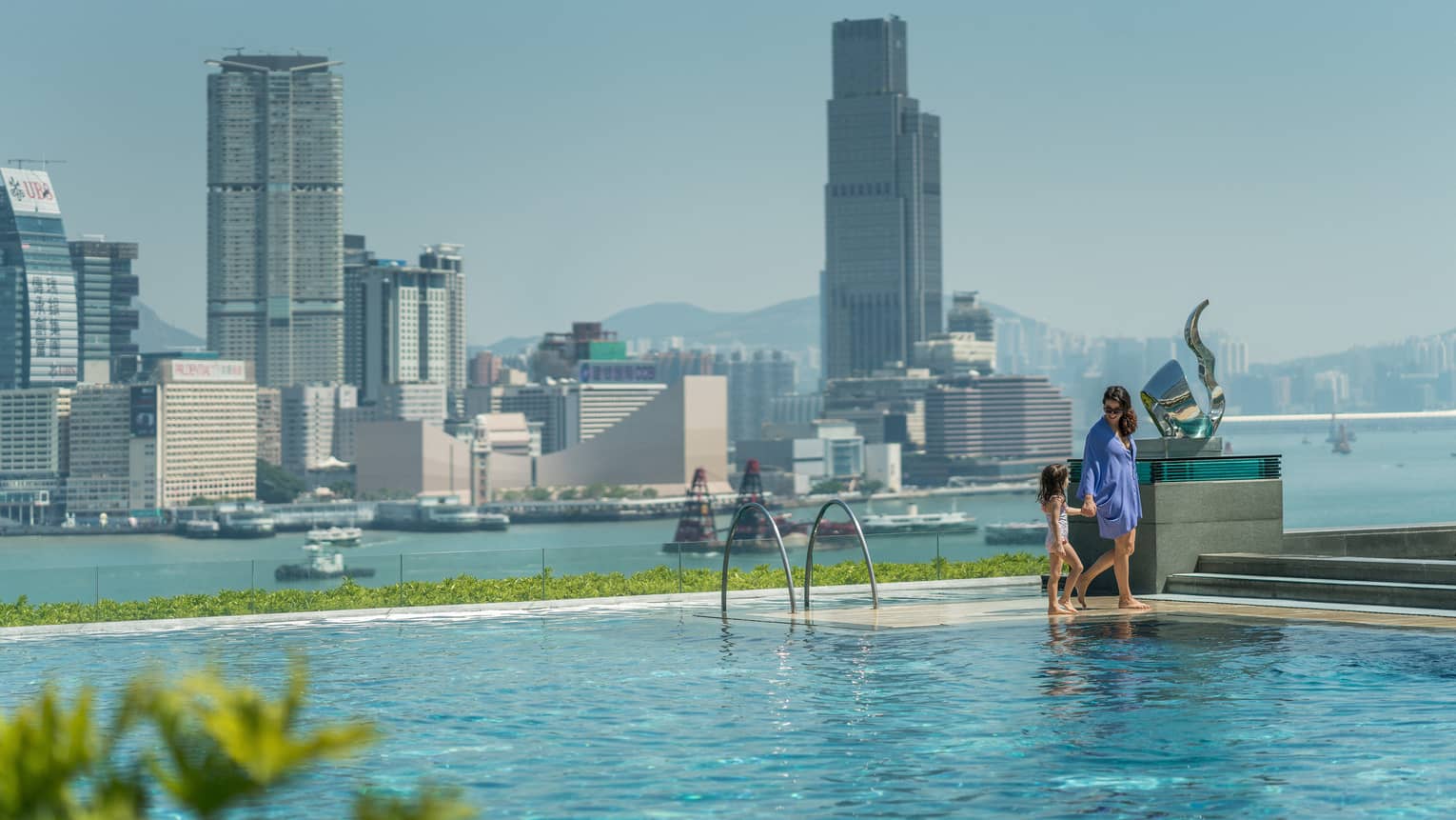 A person and child stand at the edge of a rooftop infinity pool, overlooking the harbour. The clear sky and cityscape create a serene backdrop.