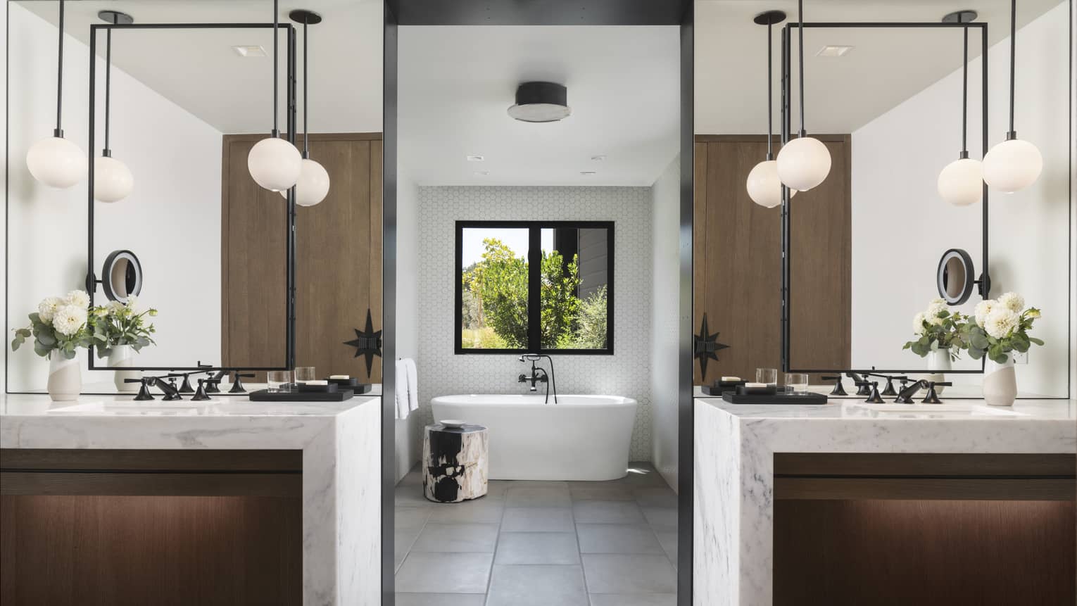Luxury resort bathroom with two vanities with marble countertops and flowers, pendant lighting, freestanding tub and window with garden view