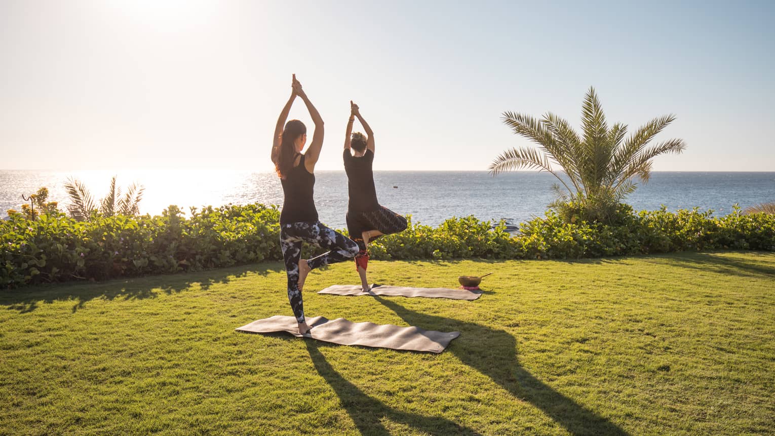 Two people enjoy yoga outdoors by the sea with the sun rising in the sky at a luxury hotel