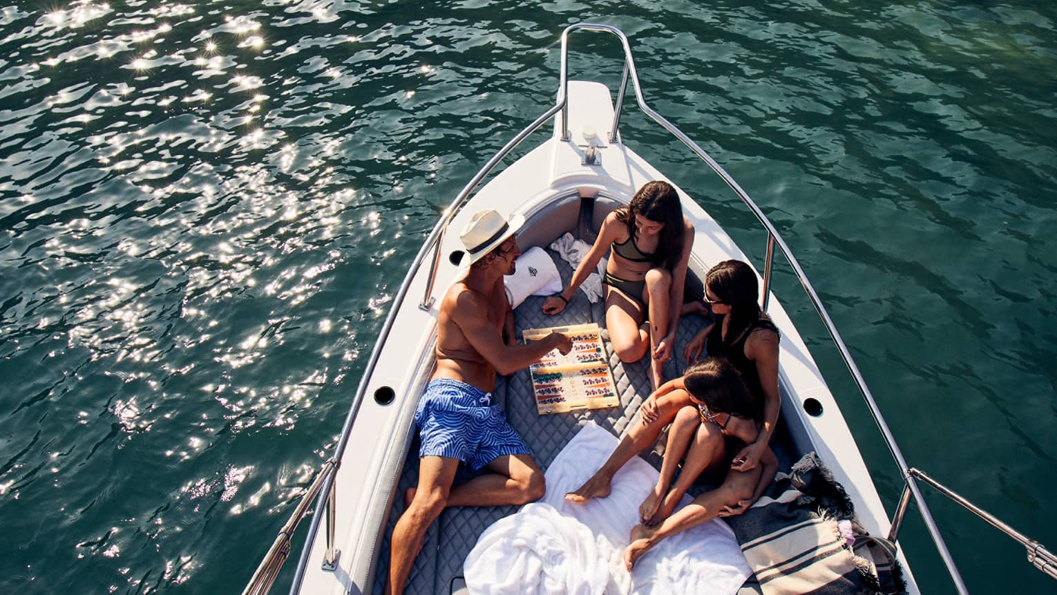 A family lounges in the bow of a luxurious boat amid towels and blankets, playing backgammon, the sun's glow on teal waters. 