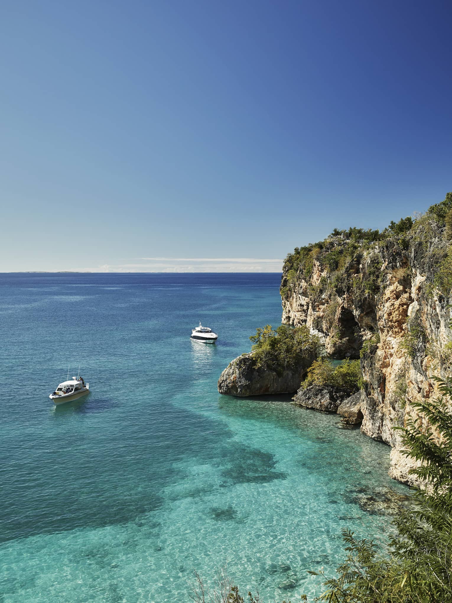 Long view of two cabin boats floating in the crystal-clear ocean beside towering rocky cliffs, a pale blue sky above.