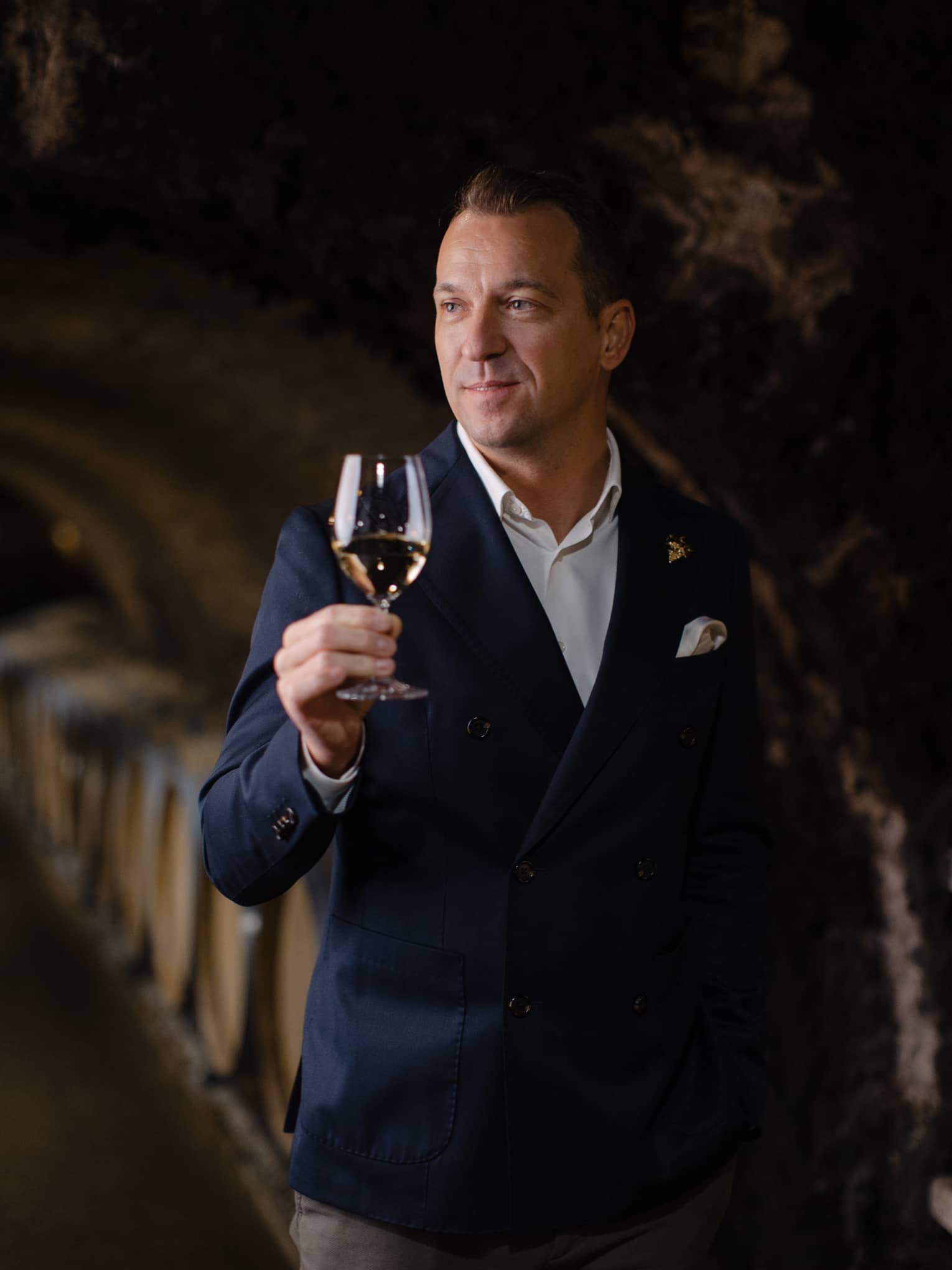 Person in a dark suit holding a glass of white wine in a wine cellar